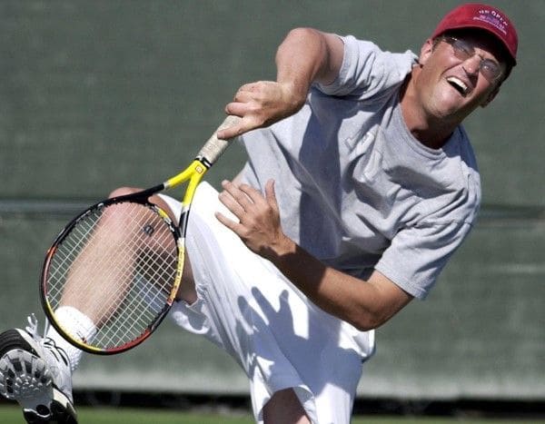 A photo of Matthew Perry making a face while hitting a tennis ball.
