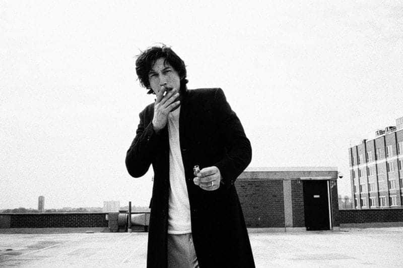Adam Driver is smoking a cigarette on a roof in a long black jacket. 