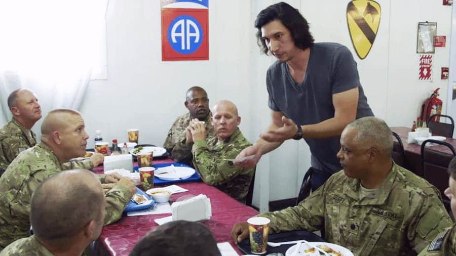 Adam Driver is talking to soldiers in uniform while they are eating a meal. 