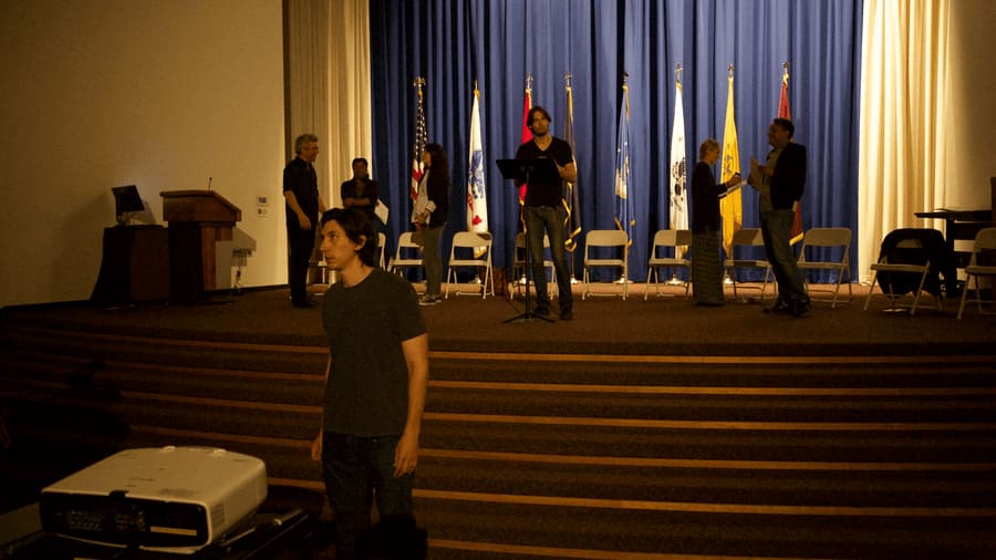 Adam Driver and fellow AITAF actors are standing around near a stage rehearsing. 