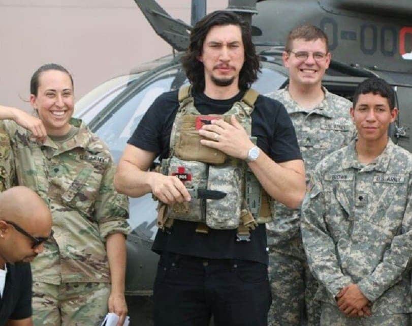 Adam Driver wearing a military vest with soldiers and a military helicopter behind them. 