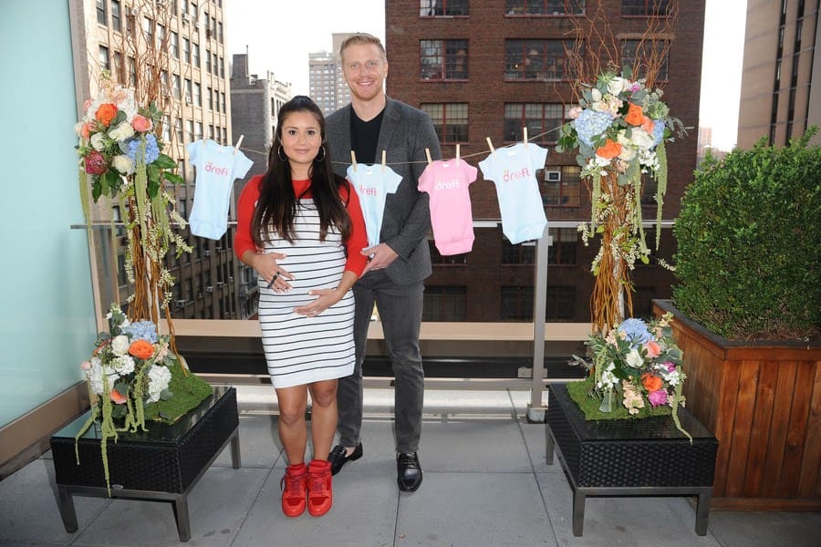 Catherine Giudici and Sean Lowe at their baby shower posing in front of baby onesies hung up on the clothesline. 