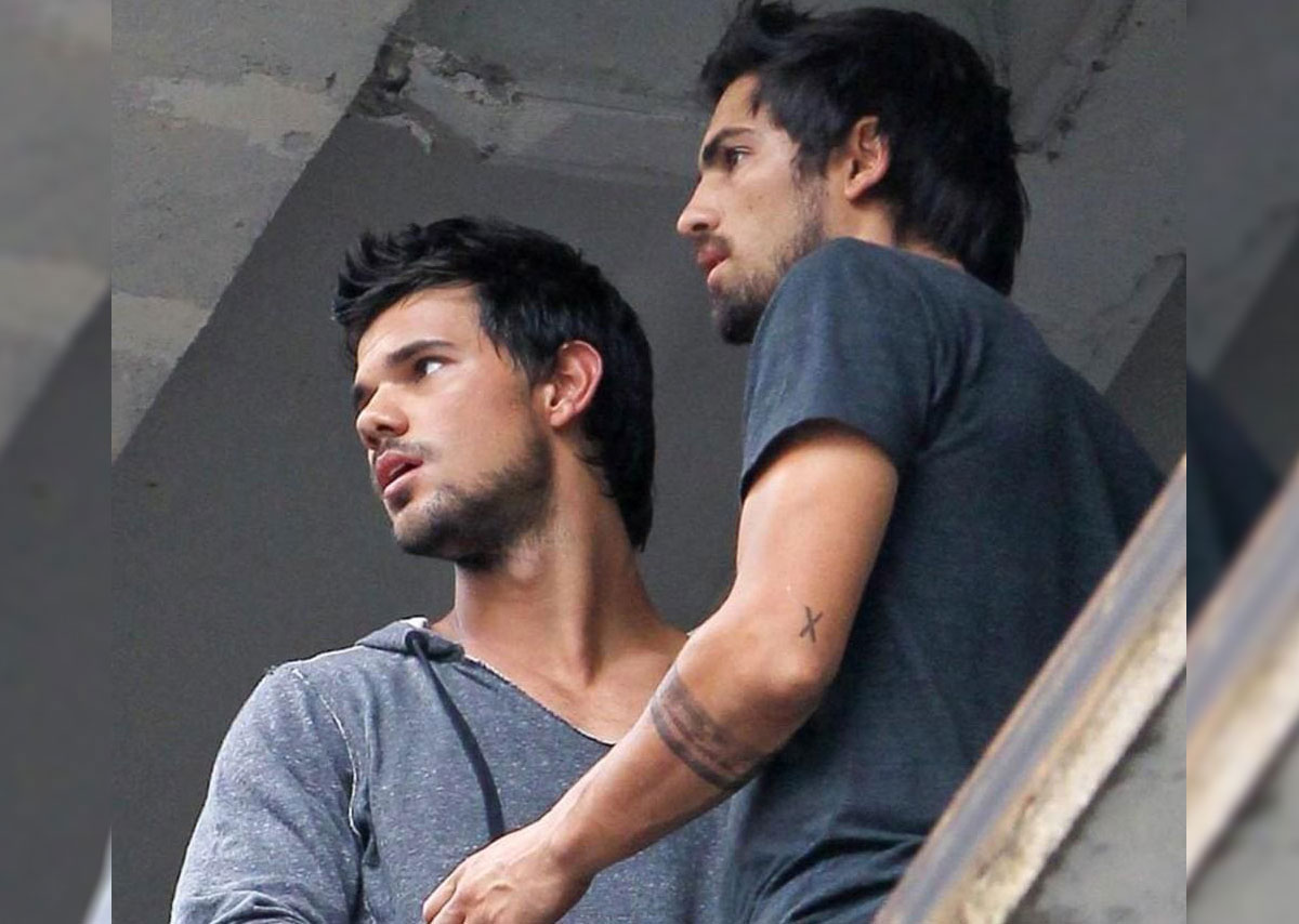 Taylor Lautner and Gabriel Nunez in matching tight short-sleeved shirts standing on a balcony.