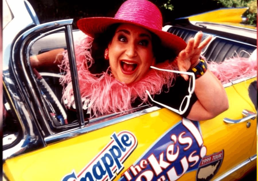 Wendy Kaufman with a pink hat, pink boa, and white sunglasses driving in a yellow Snapple car with an excited look on her face.