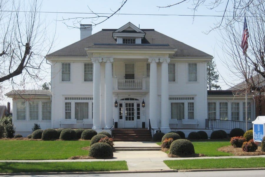 Photograph of a large white house with columns at the entrance and a few white park benches.