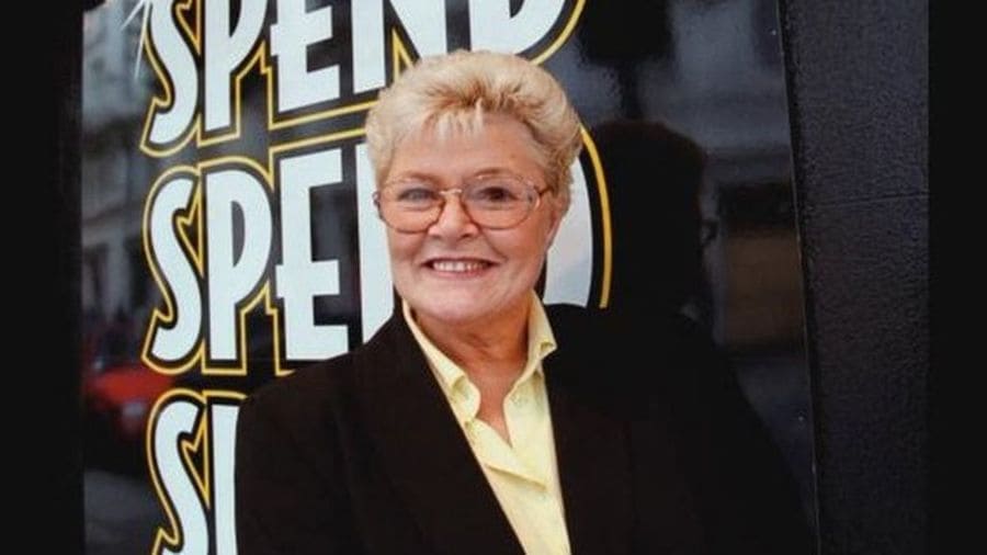 A photo of Vivian Nicholson at an older age behind a sign that says 'spend, spend, spend.'