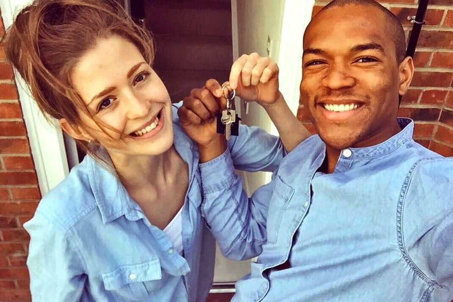 Cally and Edi were holding up the keys to their new house, standing in front of the front door and both wearing light denim shirts. 