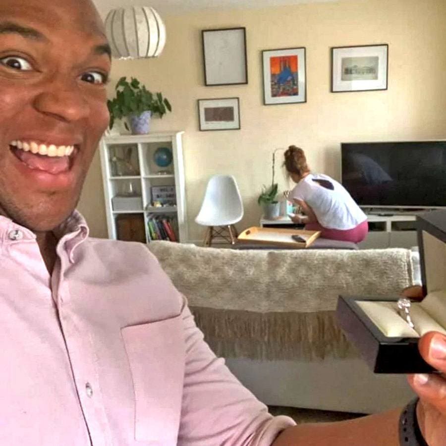 Cally is sitting next to the TV, fixing something while Edi stands behind the couch, taking an unnoticed selfie of the engagement ring. 