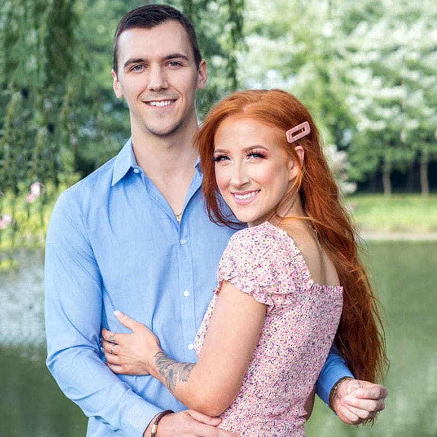 Colton and Danielle are standing near a lake in front of lots of green grass and trees while Colton holds an engagement ring behind her back. 
