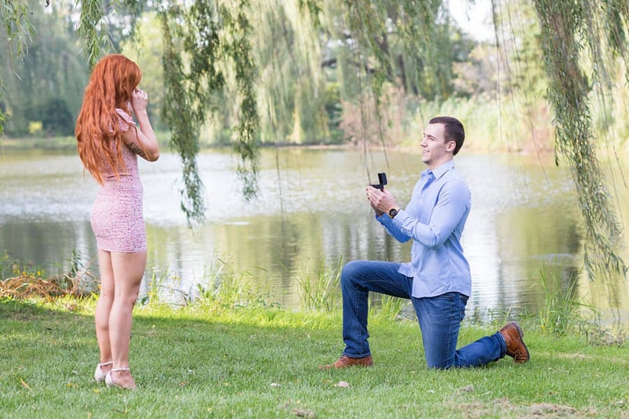 Photograph of Colton down on one knee proposing to Danielle in front of a lake with tree branches hanging over them. 