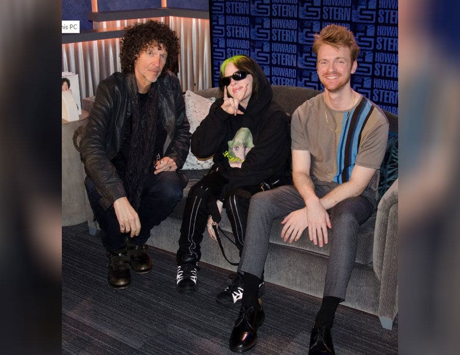 Howard Stern, Billie Eilish, and Finneas O'Connell sitting on the couch on the Howard Stern Show set.