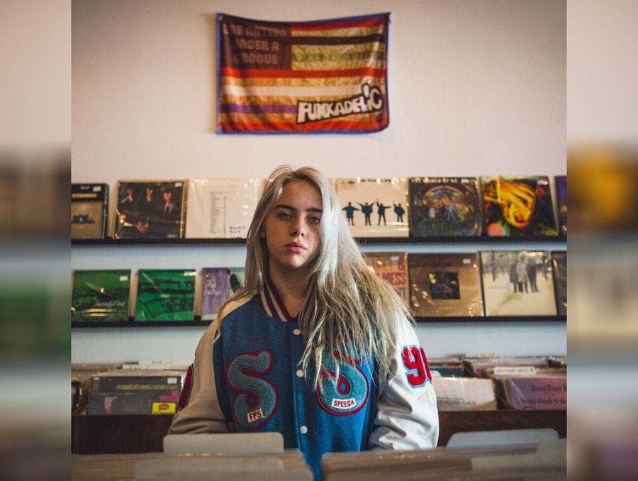 Billie Eilish is standing in a record store wearing a bomber jacket.
