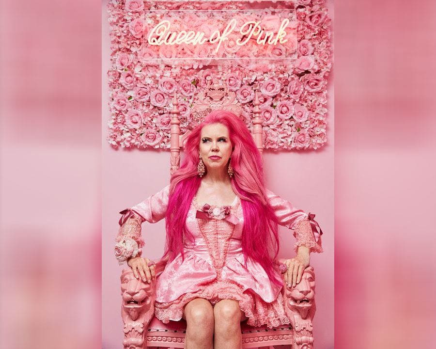 Kitten Kay Sera is sitting in her pink chair with a wall of pink flowers behind her and a pink neon sign which reads 'Queen of Pink.'
