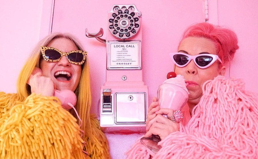 Ella London and Kitten Kay Sera are posing near a pink payphone. 