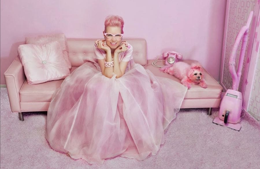 Kitten Kay Sera is sitting on her pink couch in a pink outfit with her pink dog and a telephone sitting next to her. 