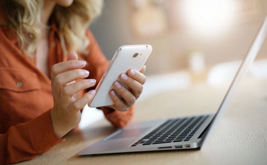 A blonde woman surfing the internet on her phone while using her laptop