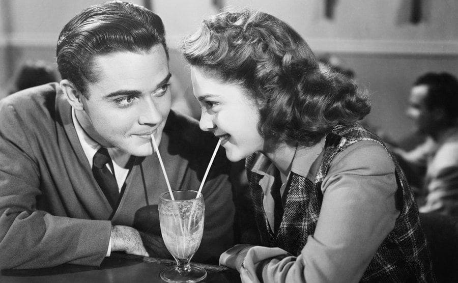 A vintage photograph of a couple drinking a milkshake with two straws while looking at each other 