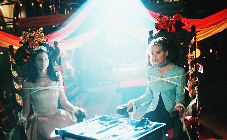 Charisma Carpenter and Sarah Michelle Gellar tied up to a large chair.