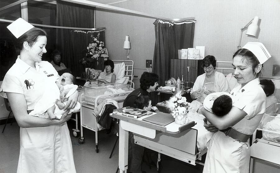A maternity ward with women in beds and nurses holding babies