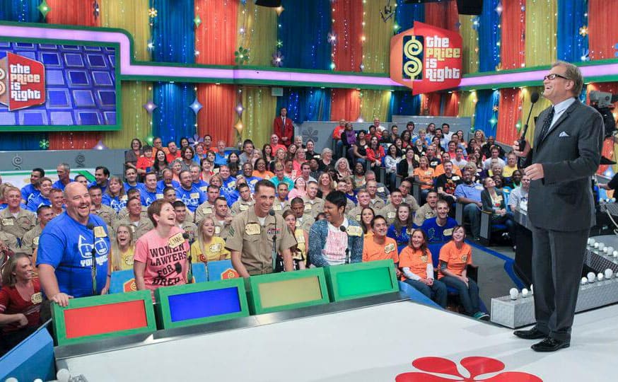 Drew Carey standing on stage with four contestants standing behind the podiums in front of the audience