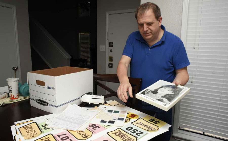Ted at his dining room table holding a photograph of one of the models from the show with his nametags and winnings sprawled out across the table
