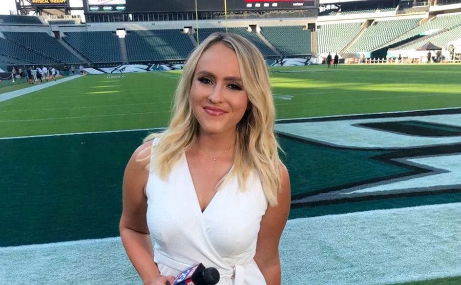 Kristen Rodgers wearing a white dress broadcasting from the field