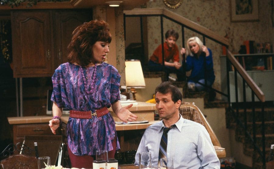 Katey Segal standing by Ed O’Neill sitting at a dining room table