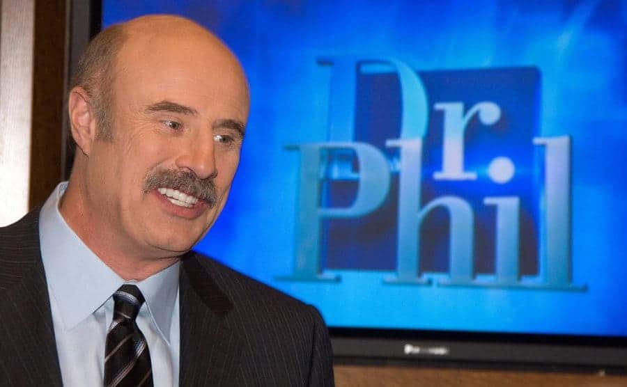 Dr Phil smiling in front of a screen with his television show logo on it