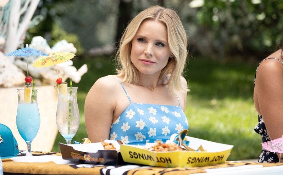 Kristen Bell sitting in front of a table with blue iced cocktails and a container of hot wings in front of her