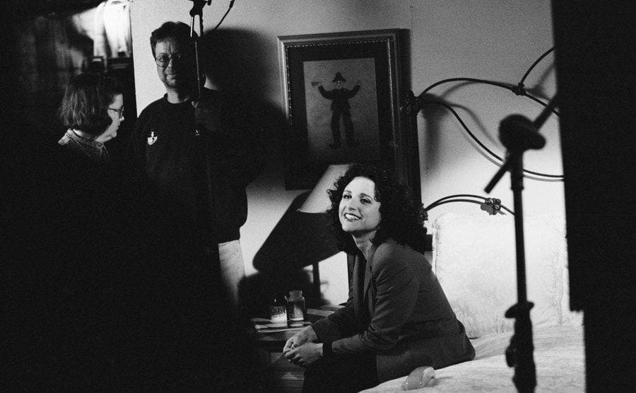 Julia Louis-Dreyfus sitting on the edge of her bed surrounded by crew on the set of Seinfeld