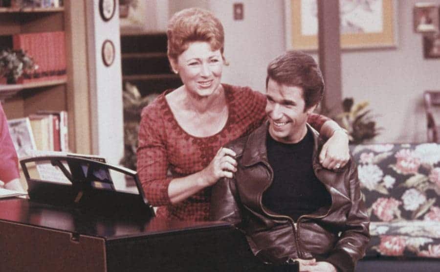 Marion Ross and Henry Winkler hugging behind a piano in a scene from happy days