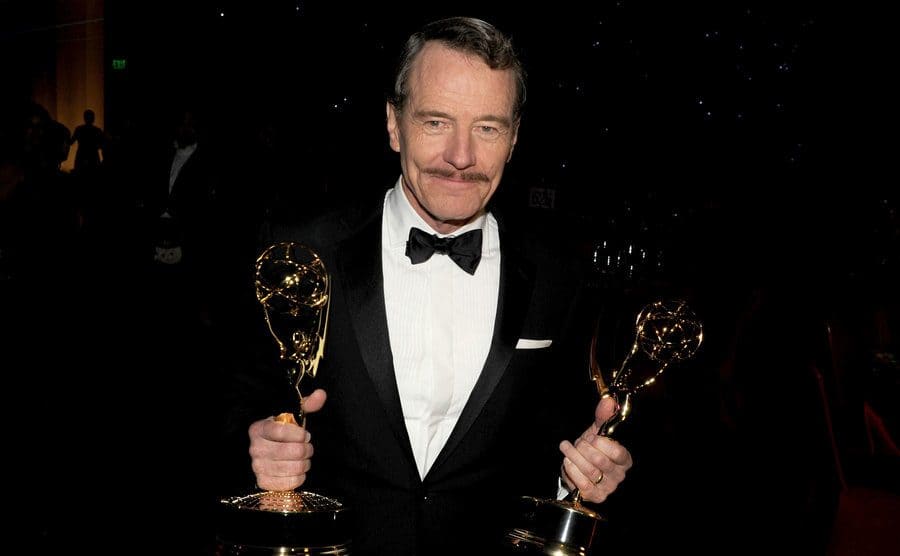 Bryan Cranston holding two Emmy Awards while wearing a tux on the red carpet