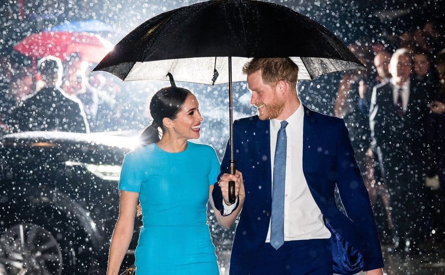 Meghan Markle and Prince Harry smiling at each other under an umbrella in the rain with a light behind them making the rain light up around them