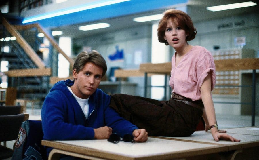 Emilio Estevez and Molly Ringwald sitting on at and on a desk in the library