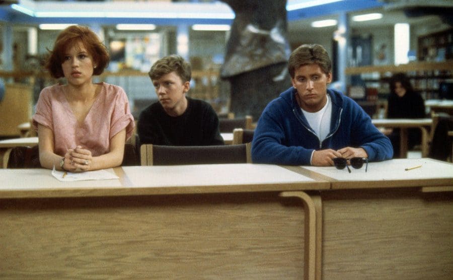 Molly Ringwald, Anthony Michael Hall, and Emilio Estevez sitting behind desks in the library
