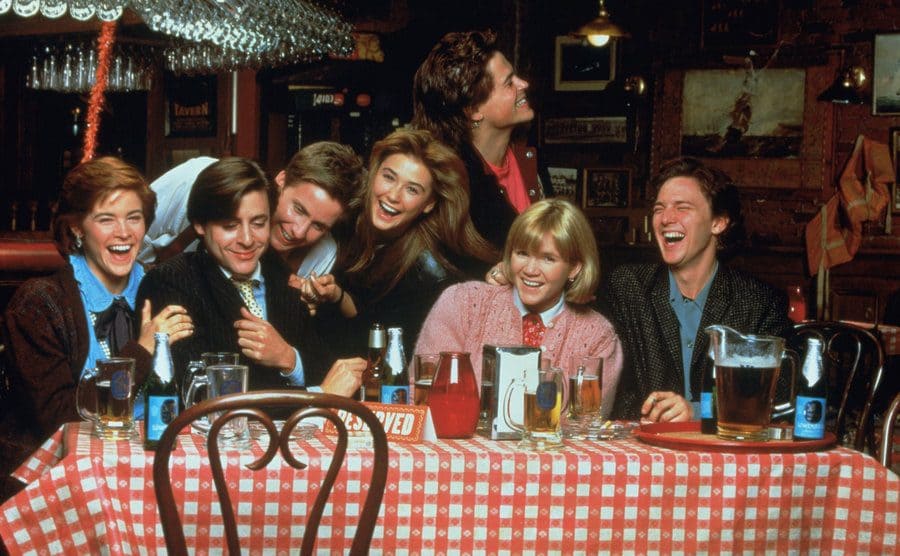 The cast of St Elmo’s Fire sitting at a bar table together, laughing