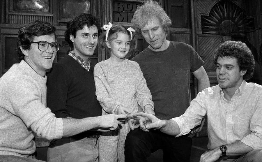 The cast of Saturday Night Live sitting with Drew Barrymore holding their pointer fingers out like in the film E.T.