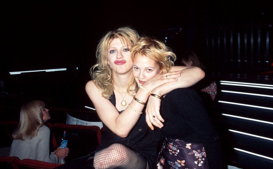 Courtney Love and Drew Barrymore sitting in a booth hugging for a photograph