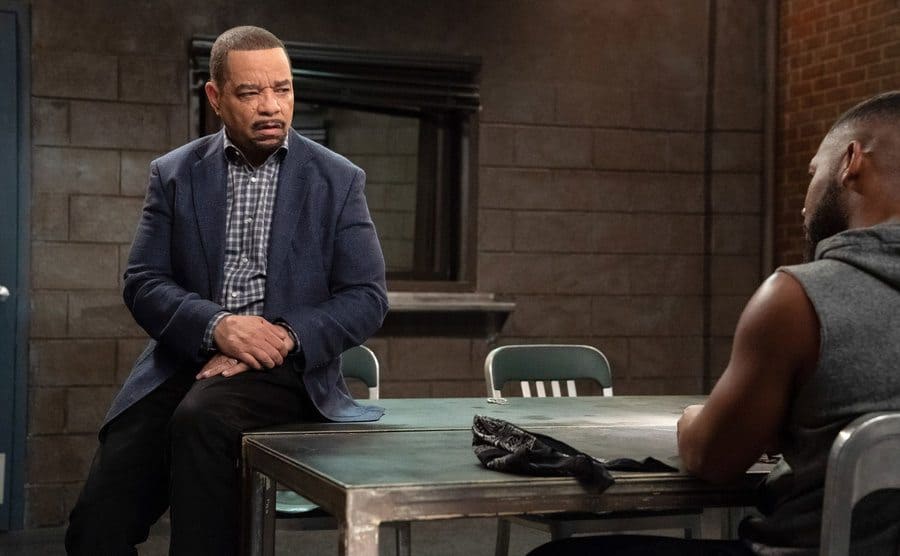 Ice T as Fin Tutuola sitting on the edge of a desk in the interrogation room with a suspect sitting across from him