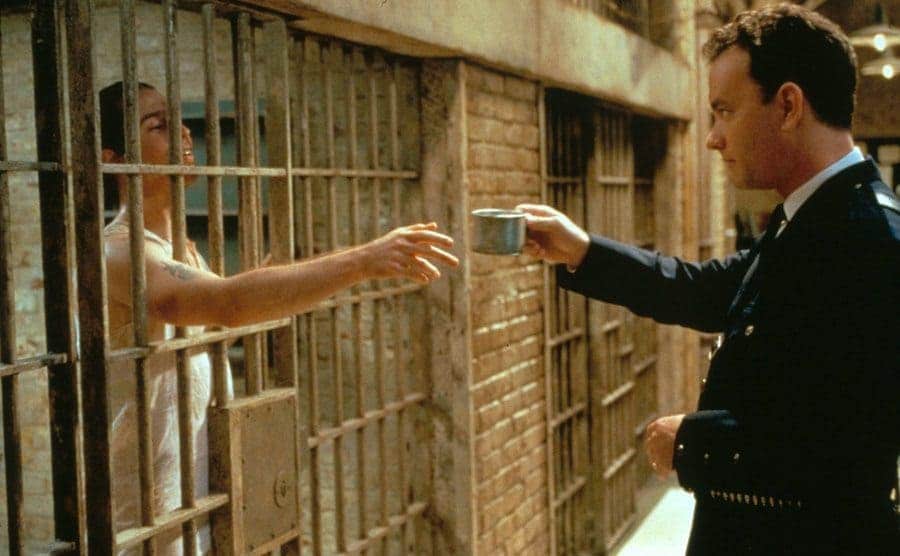 Tom Hanks passing a metal cup through the bars of a jail cell to the person inside