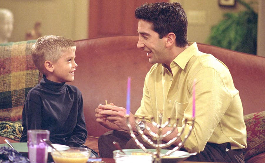 Cole Sprouse and David Schwimmer sitting on the couch with a Hanukah Menorah on the table in front of them 