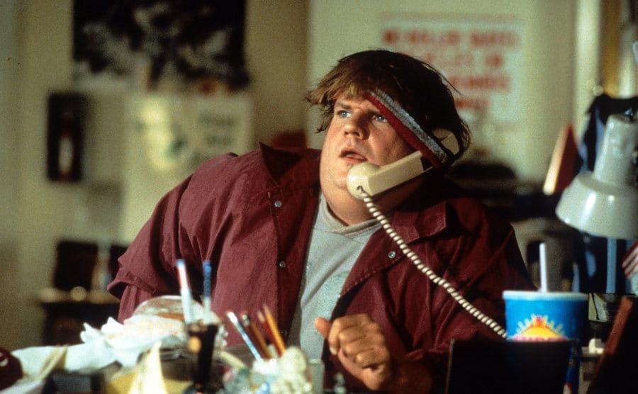 Chris Farley with a phone held onto his ear with a headband sitting behind a desk