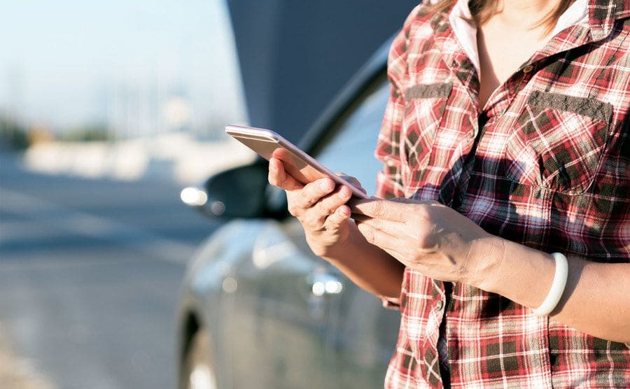 A young woman is standing beside her car looking up stuff on her phone. 