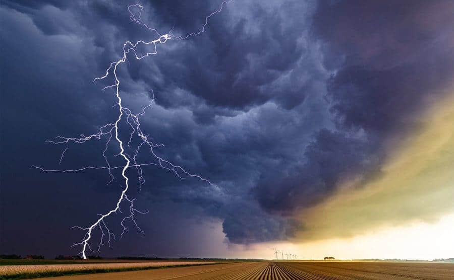A storm builds up over an uncultivated agricultural field and some wind turbines on the horizon as lightening strikes down and hits the ground. 