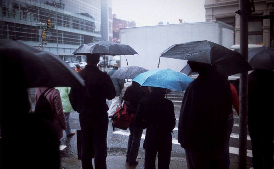 A crowd of people standing at a crosswalk in the rain. 