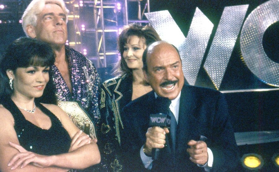 Gene Okerlund announcing with wrestlers around him 