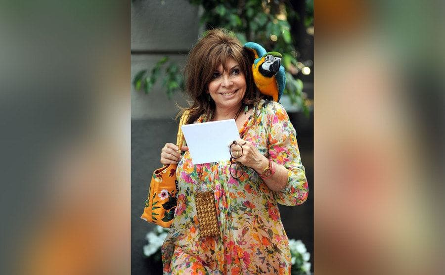 Patrizia Reggiani grinning as she walks down the street holding a piece of paper and her parrot rests on her shoulder. 