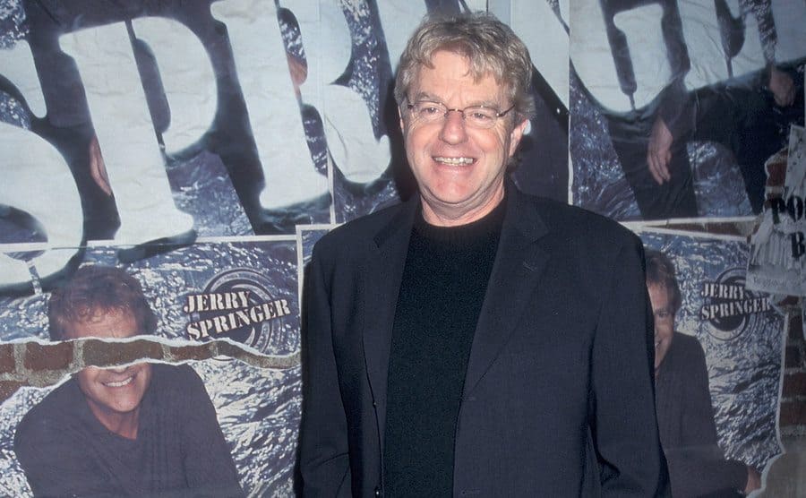 Jerry Springer posing on the set of his show with posters behind him on the wall