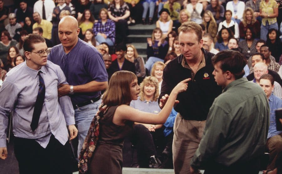 A man being held back by a security guard while a woman yells at a second man with more security standing by