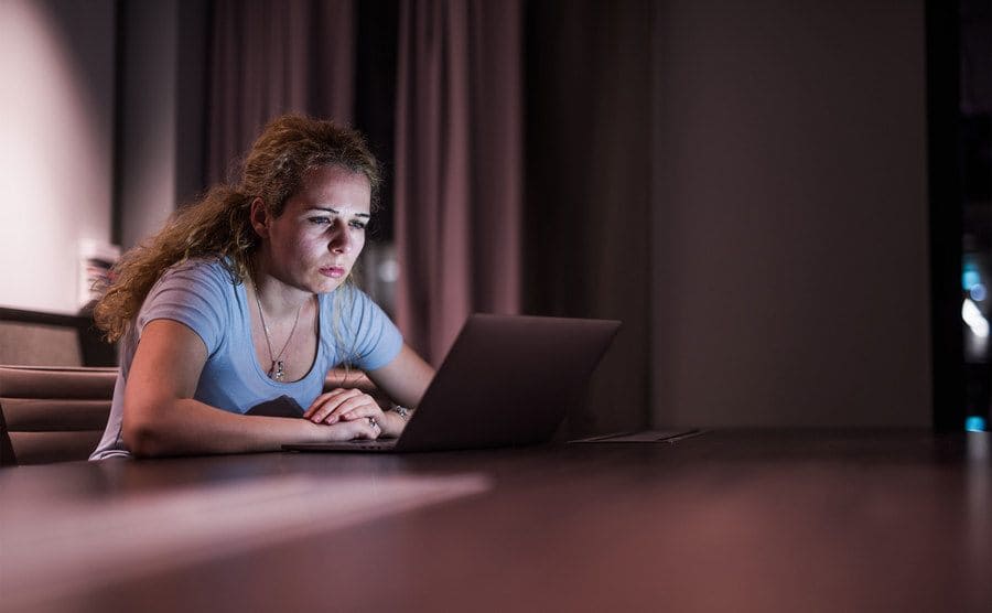 A woman is sitting at a table looking intently at her laptop screen.